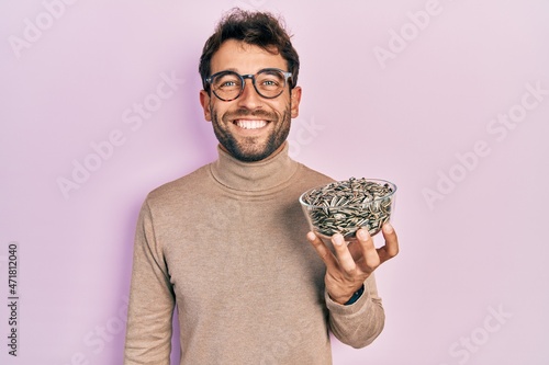Handsome man with beard football reporter holding pumpkin seeds bowl looking positive and happy standing and smiling with a confident smile showing teeth