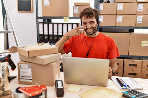 Young hispanic call center agent man working at warehouse pointing with hand finger to face and nose, smiling cheerful. beauty concept