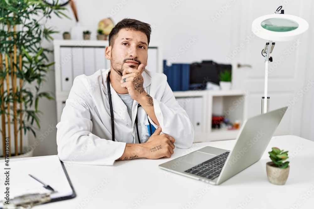 Young doctor working at the clinic using computer laptop looking confident at the camera with smile with crossed arms and hand raised on chin. thinking positive.
