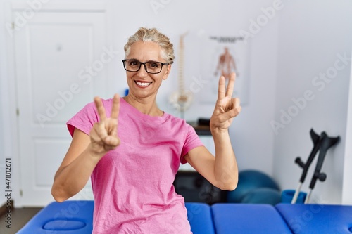 Middle age blonde woman at pain recovery clinic smiling looking to the camera showing fingers doing victory sign. number two.