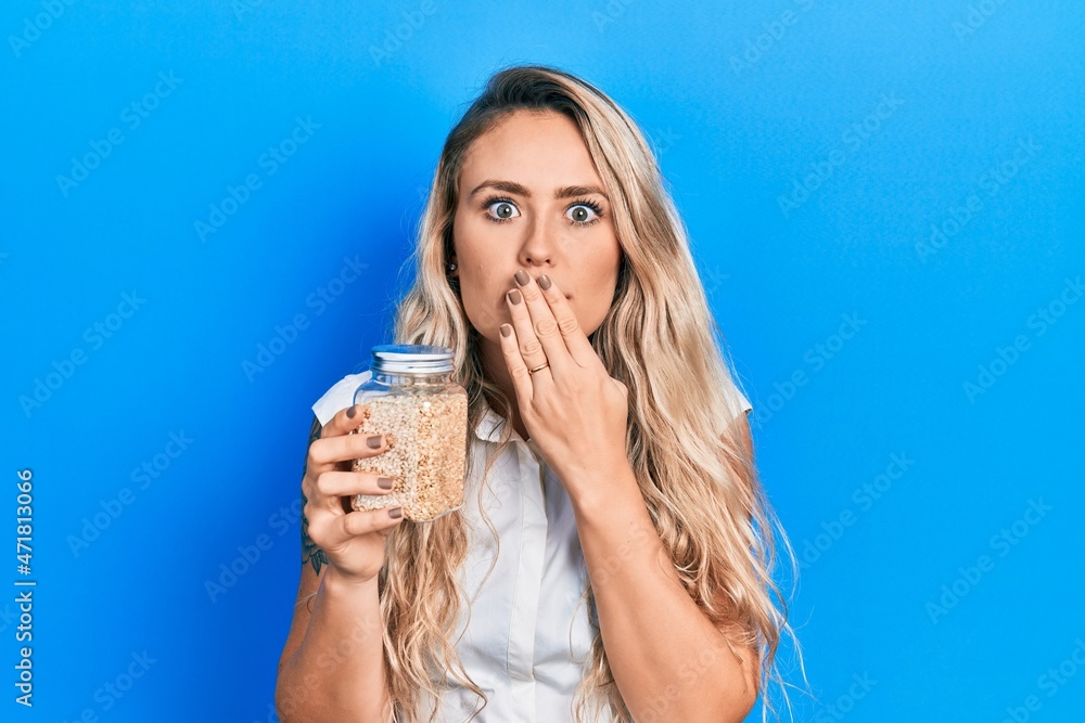 Beautiful young blonde woman holding jar with rice covering mouth with hand, shocked and afraid for mistake. surprised expression Beautiful young blonde woman holding jar with rice covering mouth with hand, shocked and afraid for mistake. surprised expression