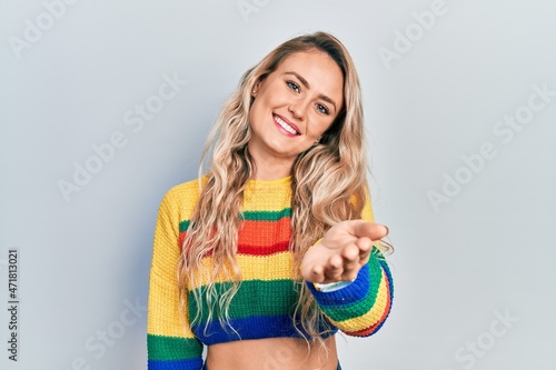 Beautiful young blonde woman wearing colored sweater smiling cheerful offering palm hand giving assistance and acceptance.