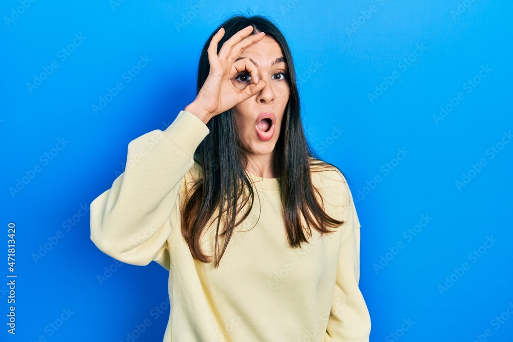 Young brunette woman wearing casual sweatshirt doing ok gesture shocked with surprised face, eye looking through fingers. unbelieving expression. Young brunette woman wearing casual sweatshirt doing ok gesture shocked with surprised face, eye looking through fingers. unbelieving expression.