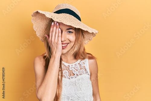 Young caucasian woman wearing summer hat covering one eye with hand, confident smile on face and surprise emotion.