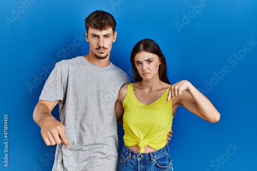 Young hispanic couple standing together over blue background pointing down looking sad and upset, indicating direction with fingers, unhappy and depressed.