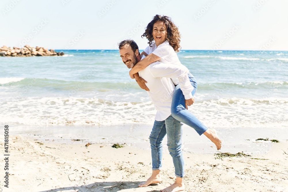 Middle age hispanic man smiling happy holding woman on his back at the beach. Middle age hispanic man smiling happy holding woman on his back at the beach.