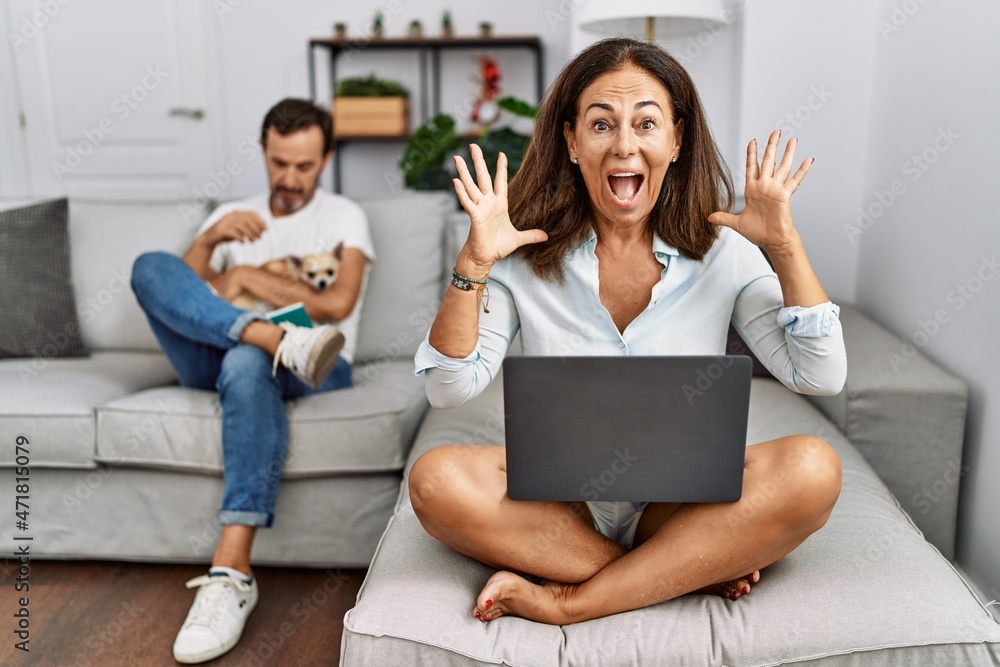 Hispanic middle age couple at home, woman using laptop celebrating crazy and amazed for success with arms raised and open eyes screaming excited. winner concept Hispanic middle age couple at home, woman using laptop celebrating crazy and amazed for success with arms raised and open eyes screaming excited. winner concept