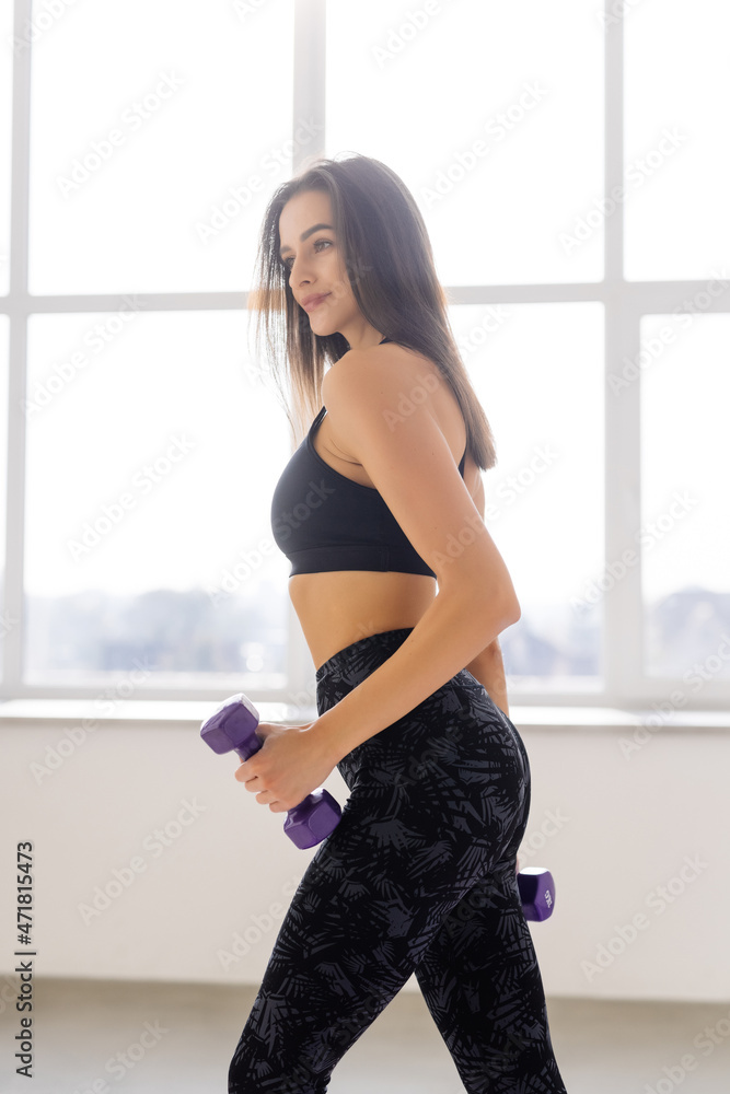 Young smiling woman exercising at home next to a window, lifting dumbbells, fitness and sports concept