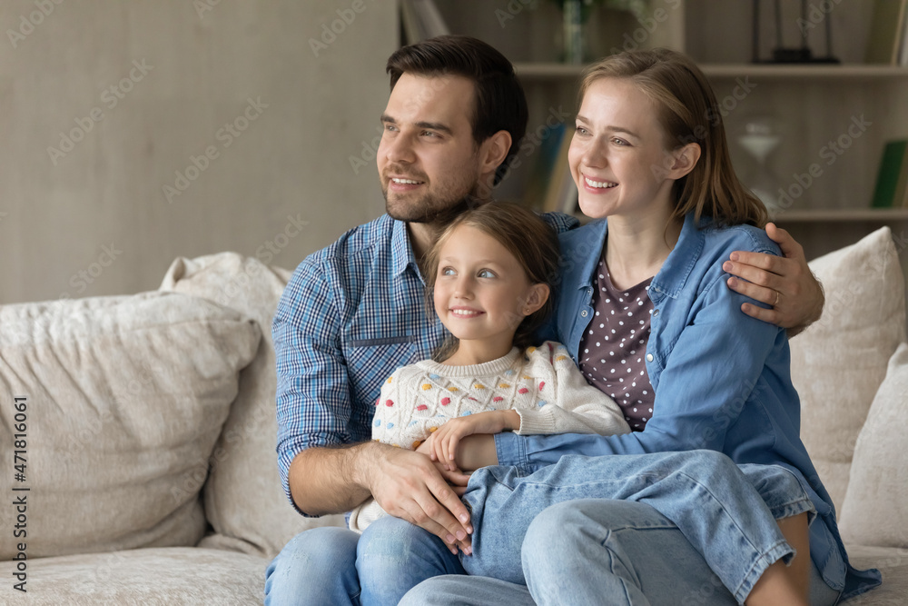 Happy loving caring young mother and father couple cuddling small child daughter looking together in distance, visualizing future or dreaming, enjoying peaceful moment at home, copy space for ad.