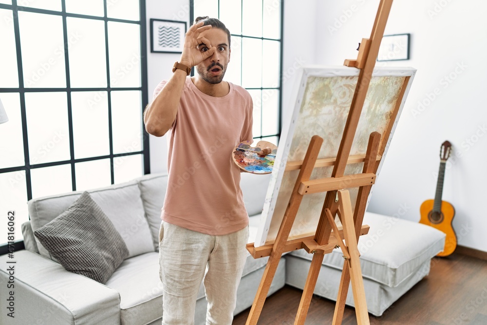 Young hispanic man with beard painting on canvas at home doing ok gesture shocked with surprised face, eye looking through fingers. unbelieving expression.