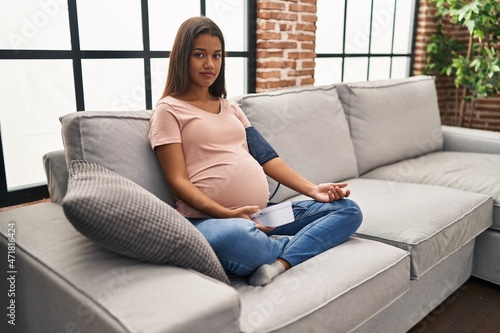 Young pregnant woman using blood pressure monitor sitting on the sofa relaxed with serious expression on face. simple and natural looking at the camera.