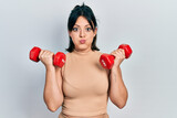 Young hispanic woman wearing sportswear using dumbbells puffing cheeks with funny face. mouth inflated with air, catching air.