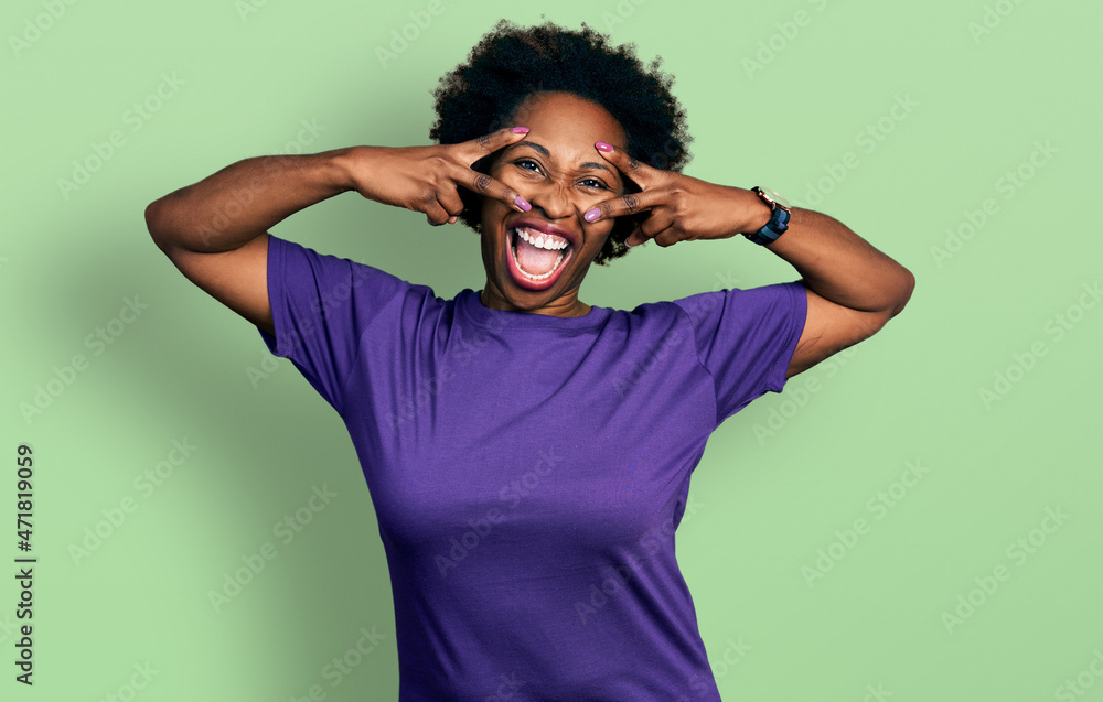 African american woman with afro hair wearing casual purple t shirt doing peace symbol with fingers over face, smiling cheerful showing victory African american woman with afro hair wearing casual purple t shirt doing peace symbol with fingers over face, smiling cheerful showing victory