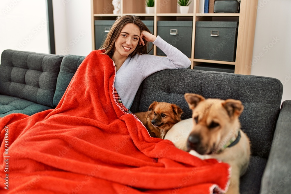 Young hispanic woman covering with blanket sitting on sofa with dogs at home
