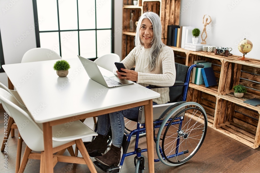 Middle age grey-haired woman using smartphone sitting on wheelchair at home Middle age grey-haired woman using smartphone sitting on wheelchair at home
