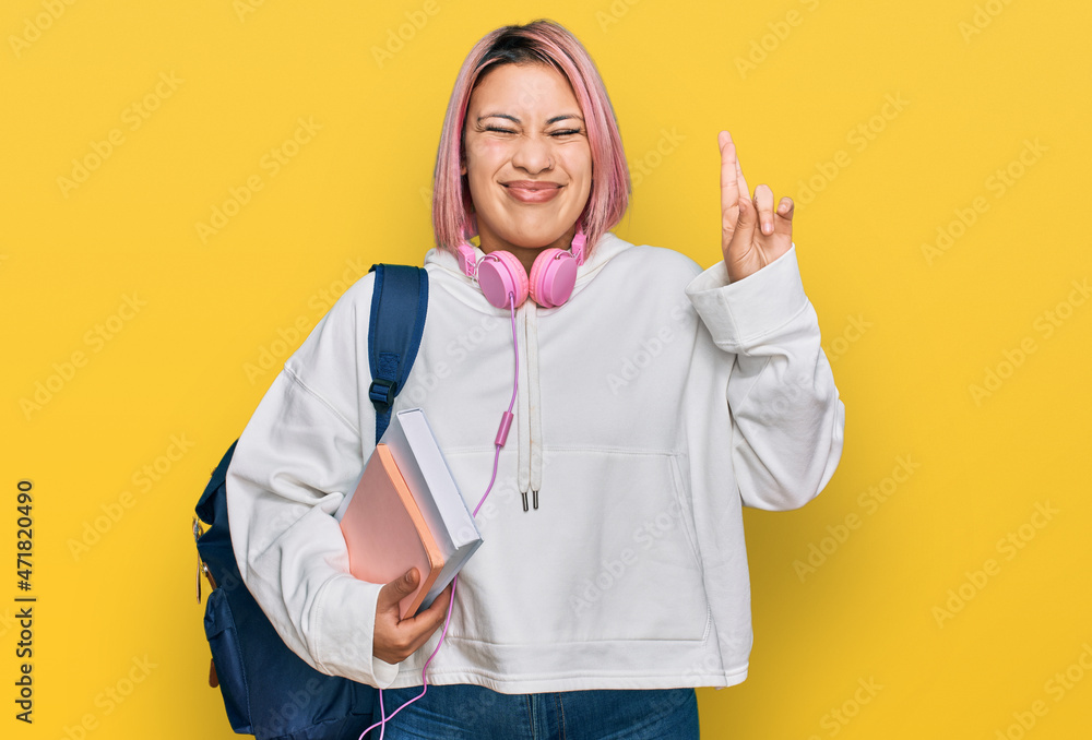 Hispanic woman with pink hair wearing student backpack and headphones gesturing finger crossed smiling with hope and eyes closed. luck and superstitious concept. Hispanic woman with pink hair wearing student backpack and headphones gesturing finger crossed smiling with hope and eyes closed. luck and superstitious concept.