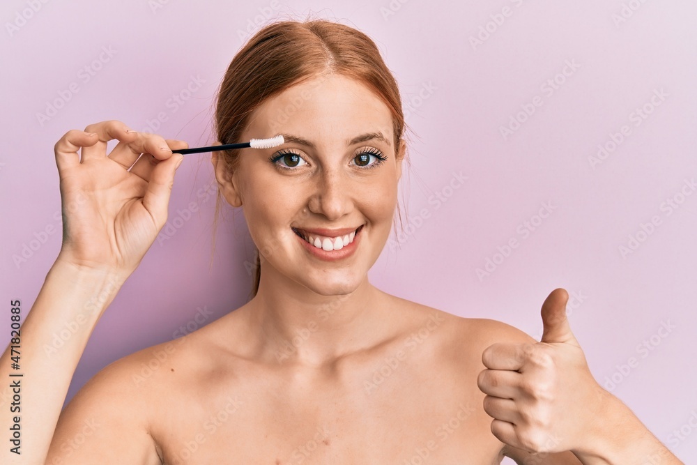 Fototapeta premium Young irish woman holding eyelash curler smiling happy and positive, thumb up doing excellent and approval sign
