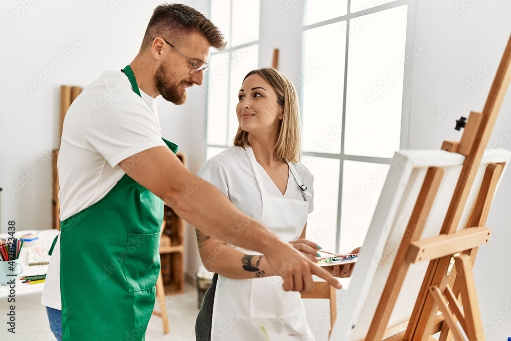 Fototapeta premium Young caucasian couple smiling happy drawing at art studio. Man standing and looking draw canvas.