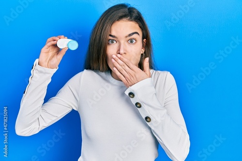 Young caucasian girl holding contact lenses covering mouth with hand, shocked and afraid for mistake. surprised expression