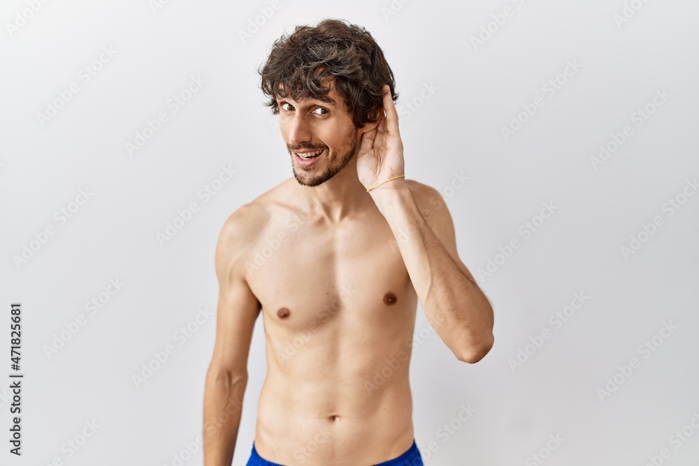 Fototapeta premium Young hispanic man standing shirtless over isolated, background smiling with hand over ear listening an hearing to rumor or gossip. deafness concept.