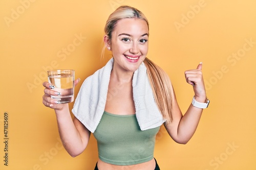 Young blonde girl wearing sportswear drinking glass of water pointing thumb up to the side smiling happy with open mouth © Krakenimages.com