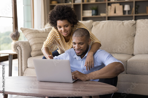 Happy young African couple focused on laptop screen watching movie, digital TV, sport match, relaxing at home. Husband and wife using computer together, pointing at screen, smiling