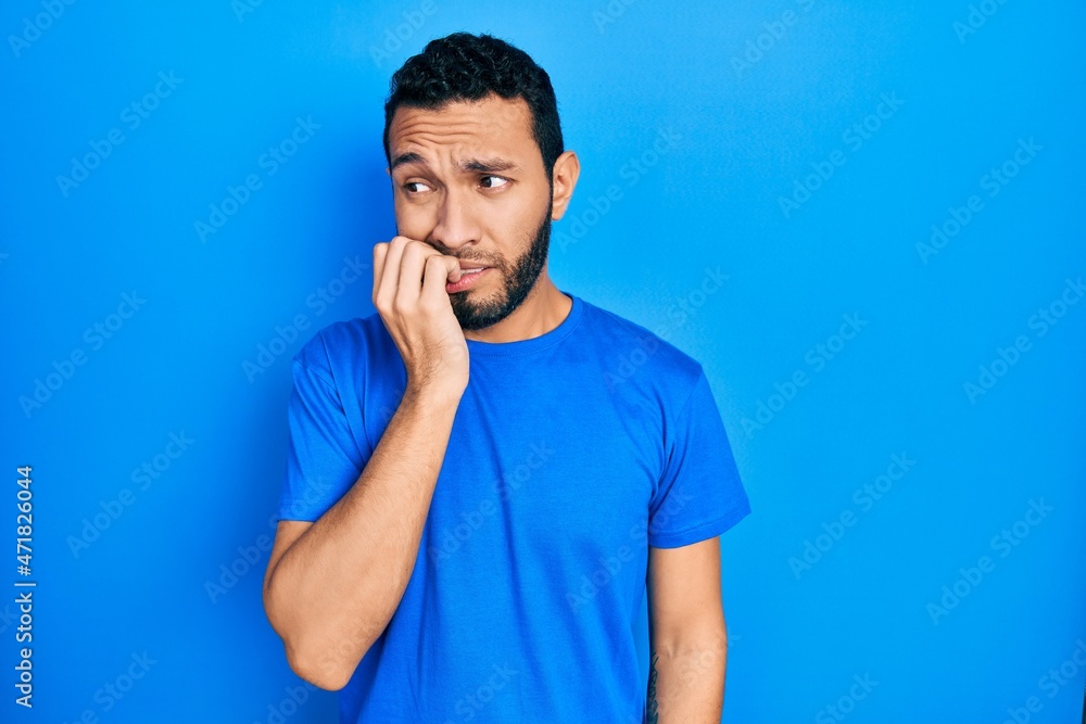 Hispanic man with beard wearing casual blue t shirt looking stressed and nervous with hands on mouth biting nails. anxiety problem. Hispanic man with beard wearing casual blue t shirt looking stressed and nervous with hands on mouth biting nails. anxiety problem.