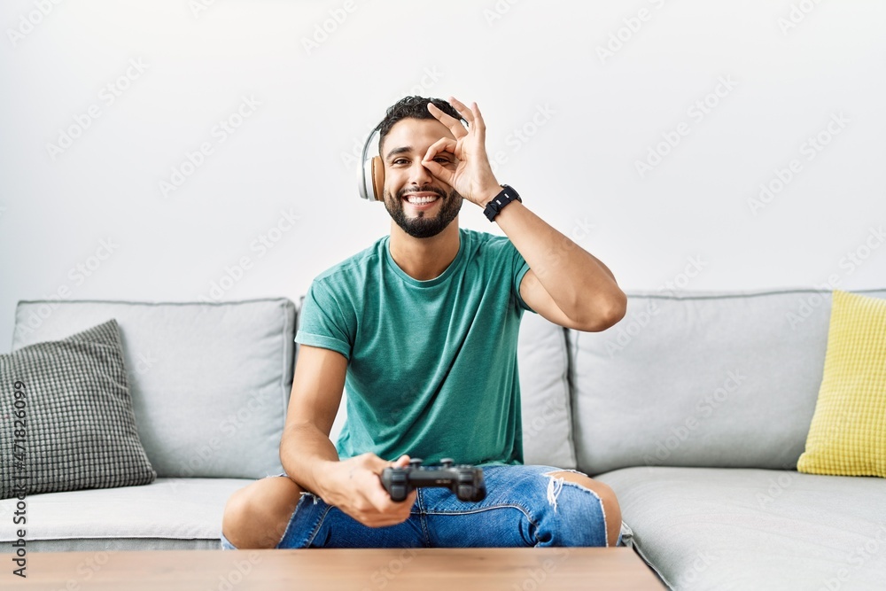 Handsome hispanic man wearing headphones playing video game holding controller smiling happy doing ok sign with hand on eye looking through fingers Handsome hispanic man wearing headphones playing video game holding controller smiling happy doing ok sign with hand on eye looking through fingers