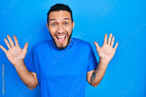 Hispanic man with beard wearing casual blue t shirt celebrating crazy and amazed for success with arms raised and open eyes screaming excited. winner concept