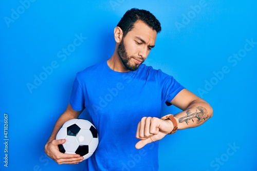 Fototapeta Naklejka Na Ścianę i Meble -  Hispanic man with beard holding soccer ball checking the time on wrist watch, relaxed and confident