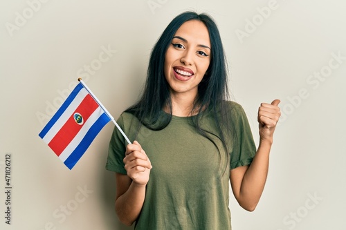 Young hispanic girl holding costa rica flag pointing thumb up to the side smiling happy with open mouth