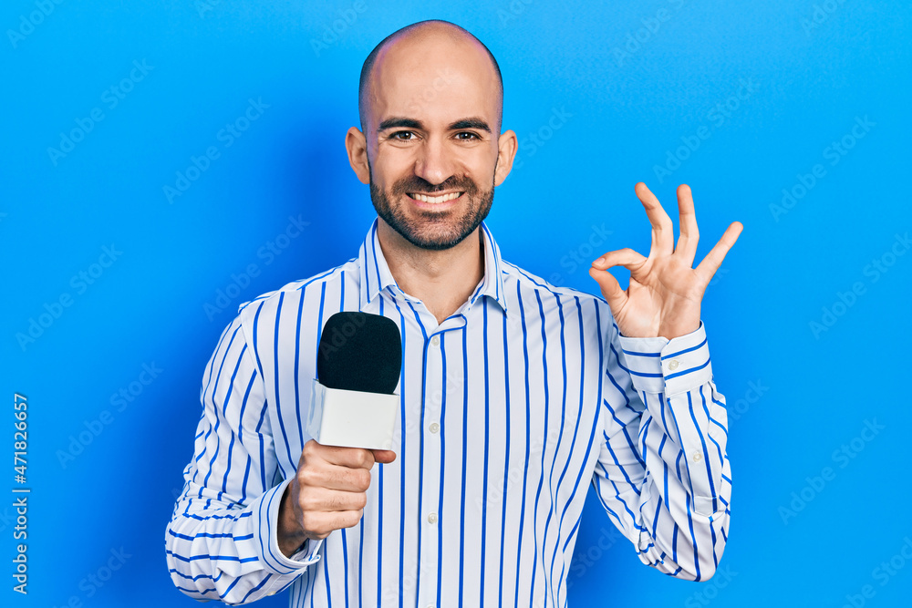 Naklejka premium Young bald man holding reporter microphone doing ok sign with fingers, smiling friendly gesturing excellent symbol