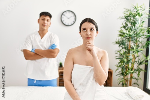 Young woman at wellness spa with professional therapist sitting on massage table serious face thinking about question with hand on chin, thoughtful about confusing idea