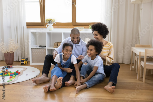 Joyful Black sibling kids and parents having fun on heating floor, tickling, giggling, laughing. Happy family couple and children playing together, enjoying leisure, activity at new home
