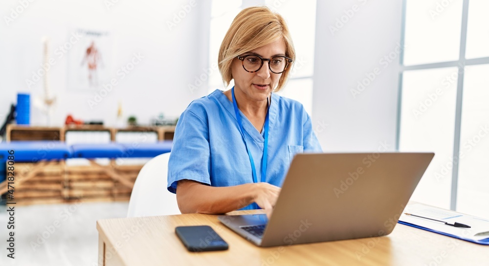 Middle age blonde woman wearing physio therapy uniform having video call at clinic Middle age blonde woman wearing physio therapy uniform having video call at clinic