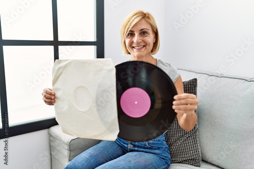 Middle age blonde woman smiling confident holding vinyl disc at home