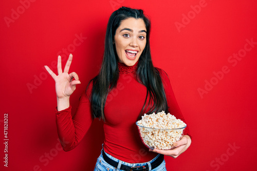Young hispanic girl eating popcorn doing ok sign with fingers  smiling friendly gesturing excellent symbol