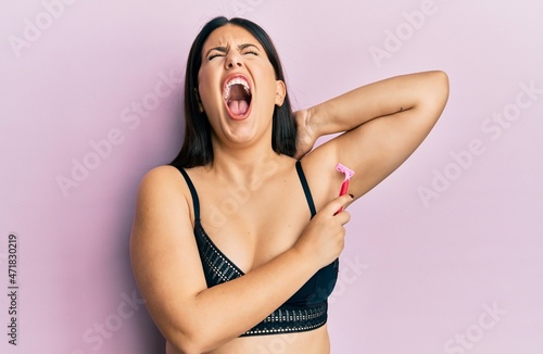 Photography Beautiful brunette woman shaving armpit hair with razor angry and mad screaming frustrated and furious, shouting with anger looking up