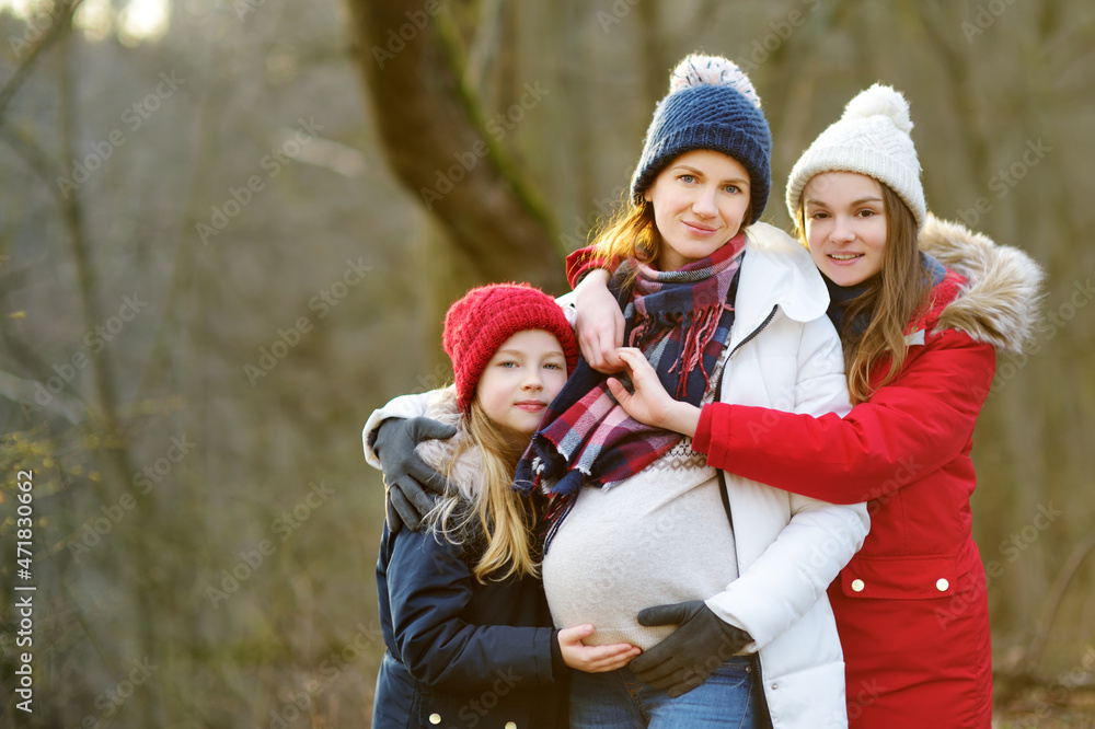 Young pregnant woman hugging her older daughters. Older siblings having fun with her pregnant mom outdoors. Mother and her kids spending time together.