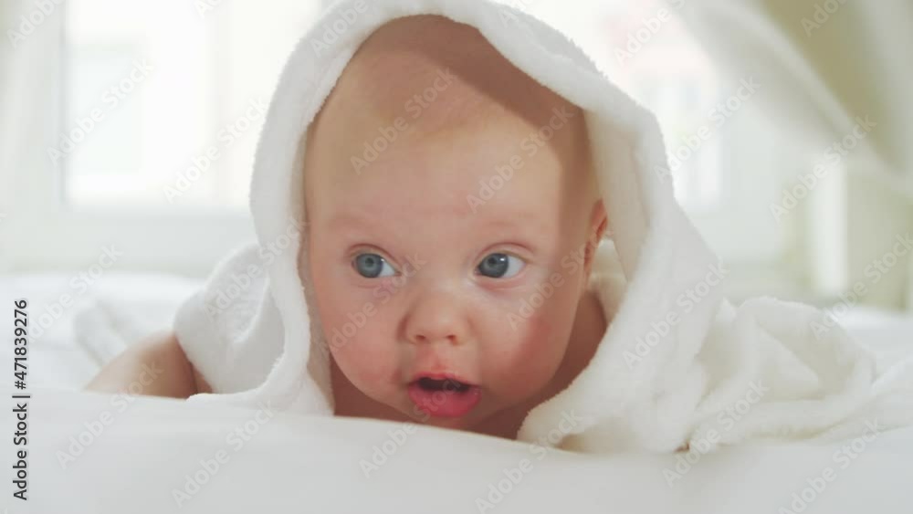 Friendly newborn baby two months old, looking into the camera lying on his stomach and covered with a white blanket, the child barely holds his head and smiles. Baby is lying on his stomach with Friendly newborn baby two months old, looking into the camera lying on his stomach and covered with a white blanket, the child barely holds his head and smiles. Baby is lying on his stomach with