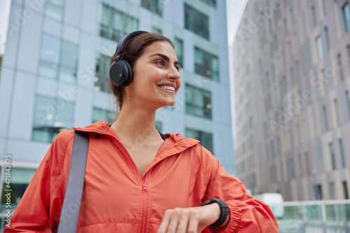 Outdoor shot of happy satisfied woman in anorak glad to monitor fitness results on smartwatch listens music from playlist has glad expression poses against blurred background. Sporty lifestyle