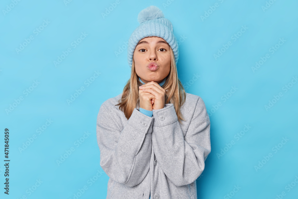 Lovely good looking young woman keeps hands under chin folds lips being sensual and cute expresses love wears hat and grey jacket isolated over blue background blows air kiss waits for mwah. Lovely good looking young woman keeps hands under chin folds lips being sensual and cute expresses love wears hat and grey jacket isolated over blue background blows air kiss waits for mwah.