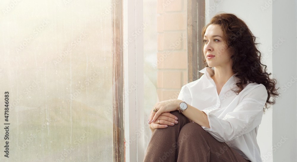 A young beautiful woman in a white shirt sits with a pensive view on the windowsill and looks out the window. The concept of sadness, longing, unrequited love, separation from a loved one. Copy space A young beautiful woman in a white shirt sits with a pensive view on the windowsill and looks out the window. The concept of sadness, longing, unrequited love, separation from a loved one. Copy space