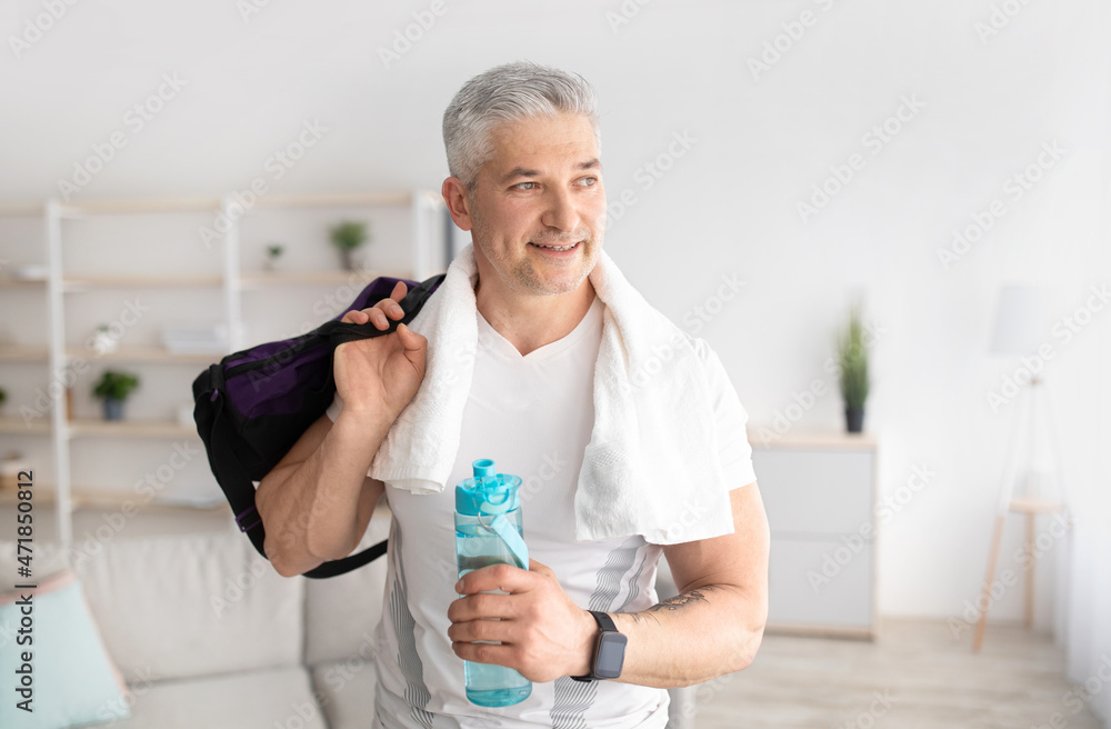 Fototapeta premium Sporty mature man holding bottle of water and sports bag, posing after domestic training and smiling, free space