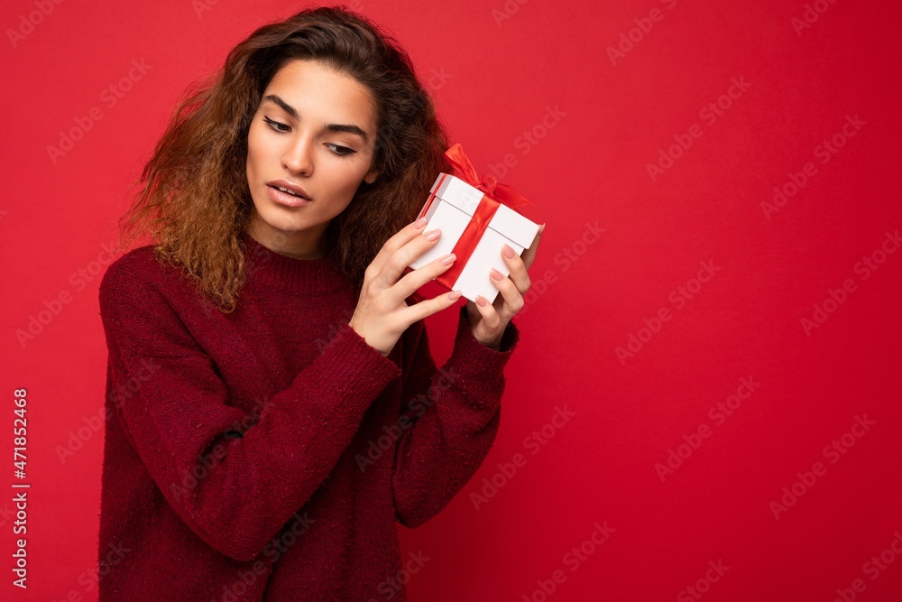 Photo of beautiful attractive charming young brunette curly woman wearing stylish dark red sweater isolated over red background wall with free space for text and holding white gift box with red ribbon Photo of beautiful attractive charming young brunette curly woman wearing stylish dark red sweater isolated over red background wall with free space for text and holding white gift box with red ribbon