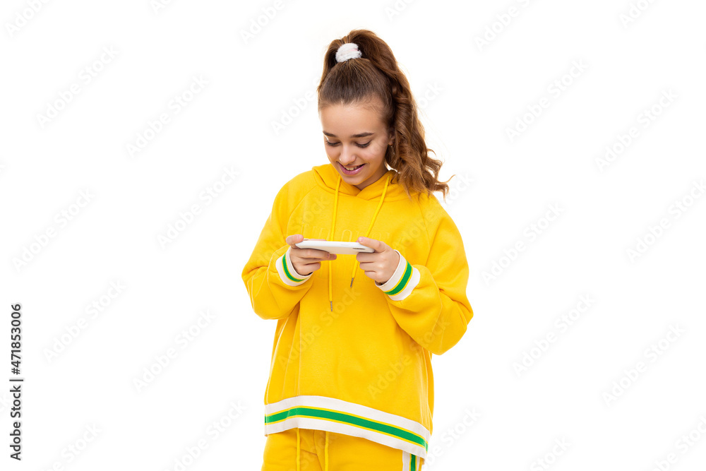 Pretty beautiful smiling cheerful cute girl with dark hair with a ponytail in a stylish yellow tracksuit with green stripes plays a mobile phone with a white case isolated on a white background with Pretty beautiful smiling cheerful cute girl with dark hair with a ponytail in a stylish yellow tracksuit with green stripes plays a mobile phone with a white case isolated on a white background with