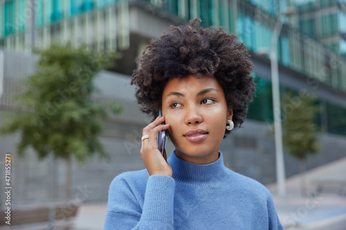 Photo of nice looking woman receives phone call in roaming talks with best friend satisfie with good mobile connection dressed casually poses outside against blurred background. Communication