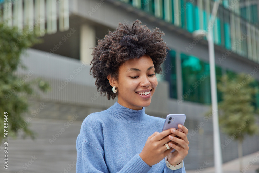 Half length shot of positive glad woman in blue turtleneck poses in urbanity with mobile phone explores new city downloads application has chat conversation. People technology and free time. - obrazy, fototapety, plakaty 