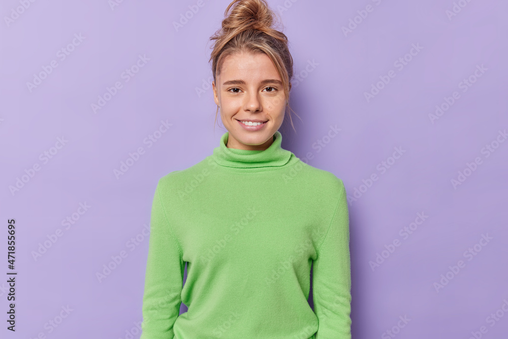 Portrait of satisfied millennial girl smiles gently has healthy skin no makeup natural beauty looks directly at camera with satisfied expression wears green poloneck isolated over purple background Portrait of satisfied millennial girl smiles gently has healthy skin no makeup natural beauty looks directly at camera with satisfied expression wears green poloneck isolated over purple background