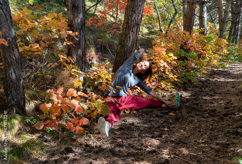 Smiling woman with red Indian pants sitting in the forest under the trees on the path with autumn foliage, yoga poses 
